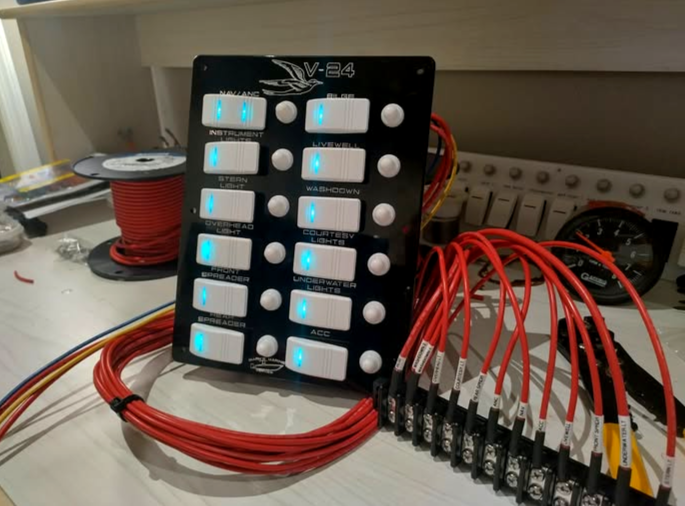 Marine switch panel labeled V-24 with illuminated toggle switches and a bundle of red wiring harness leads on a workbench.