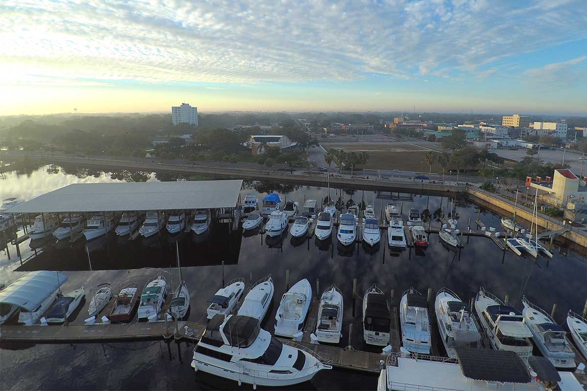Aerial view of boats docked at a marina with a covered boat shelter and city buildings beyond, at sunrise.