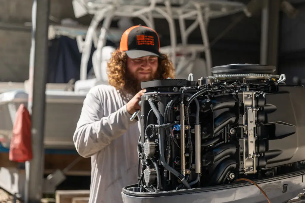 Technician working on an outboard motor at a boatyard, with a boat and storage racks in the background.
