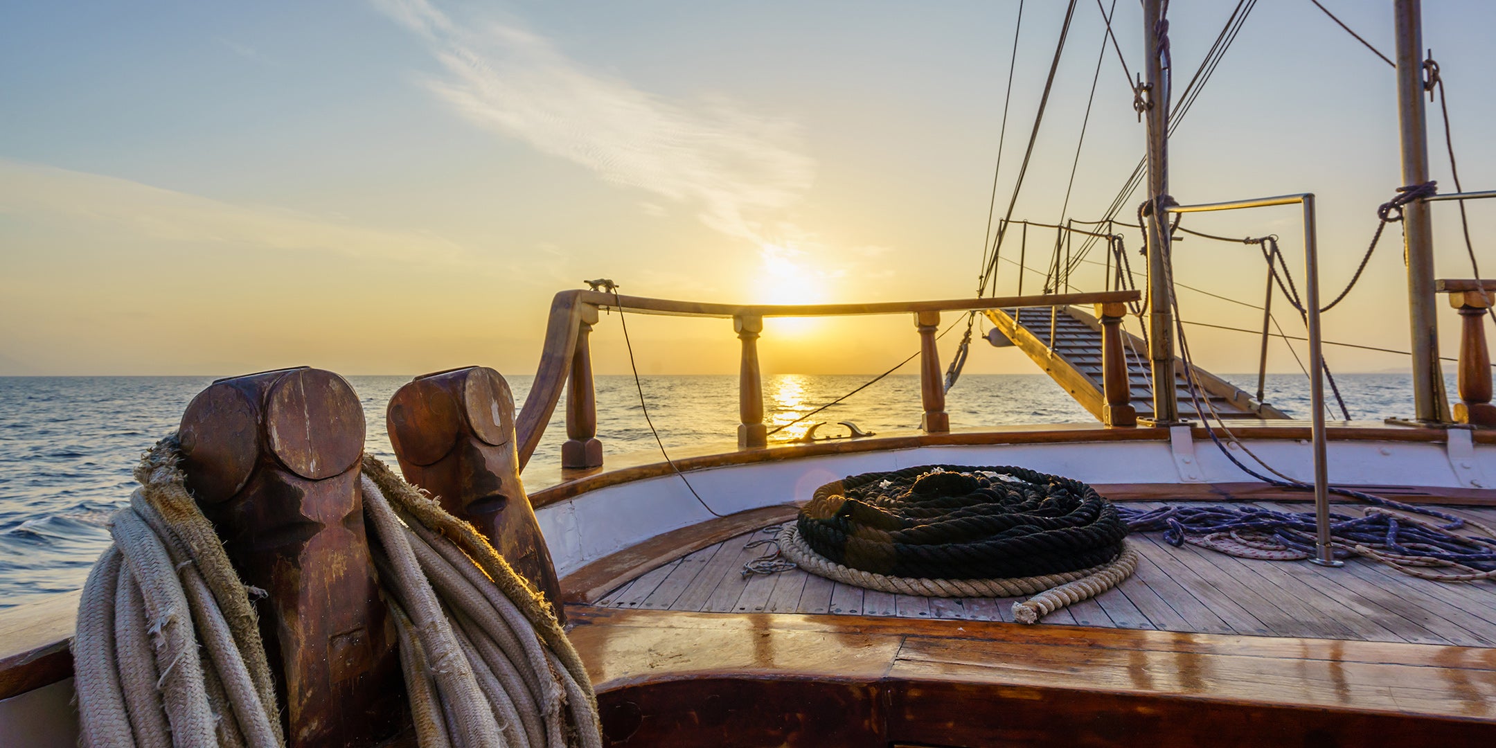 Wooden sailing yacht deck with coiled rigging lines and a teak railing at sunset over calm open water.