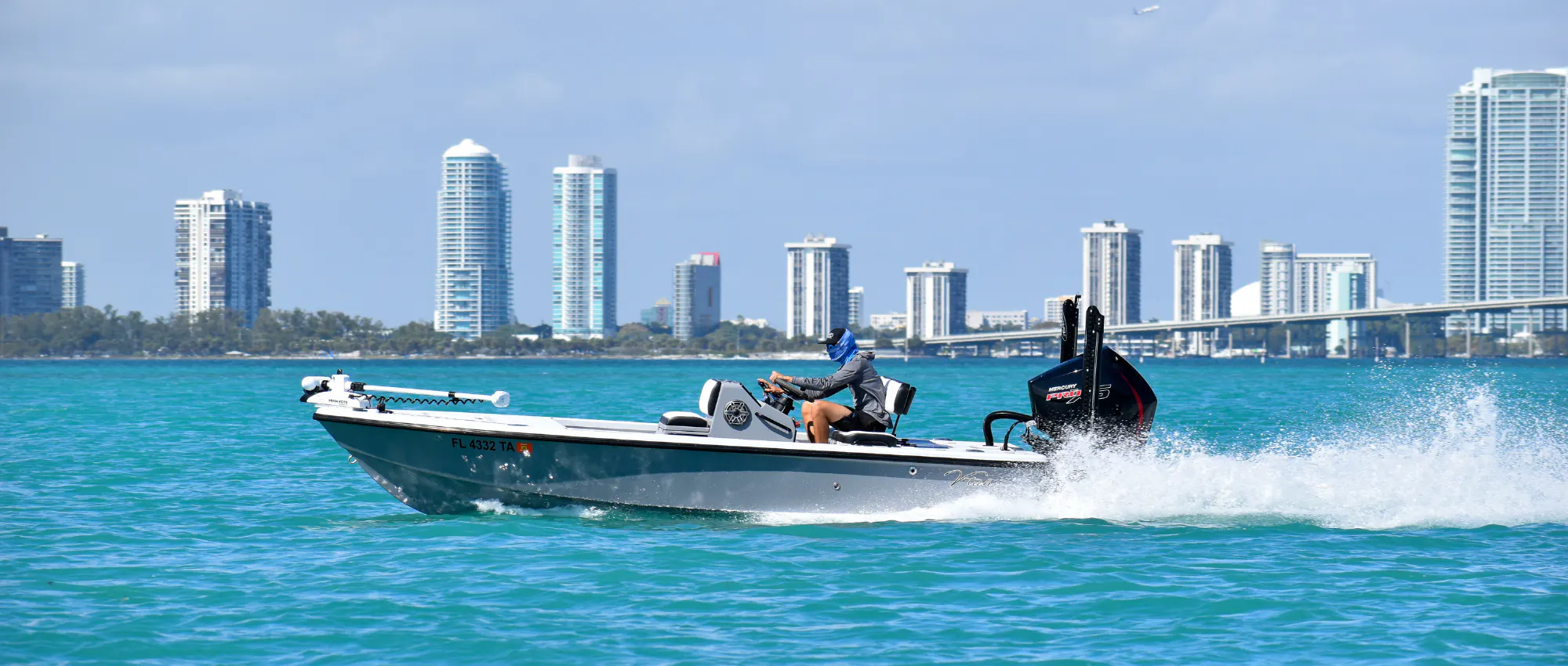 Flats boat with a Mercury outboard running across turquoise bay water in front of a high-rise skyline, Miami area.