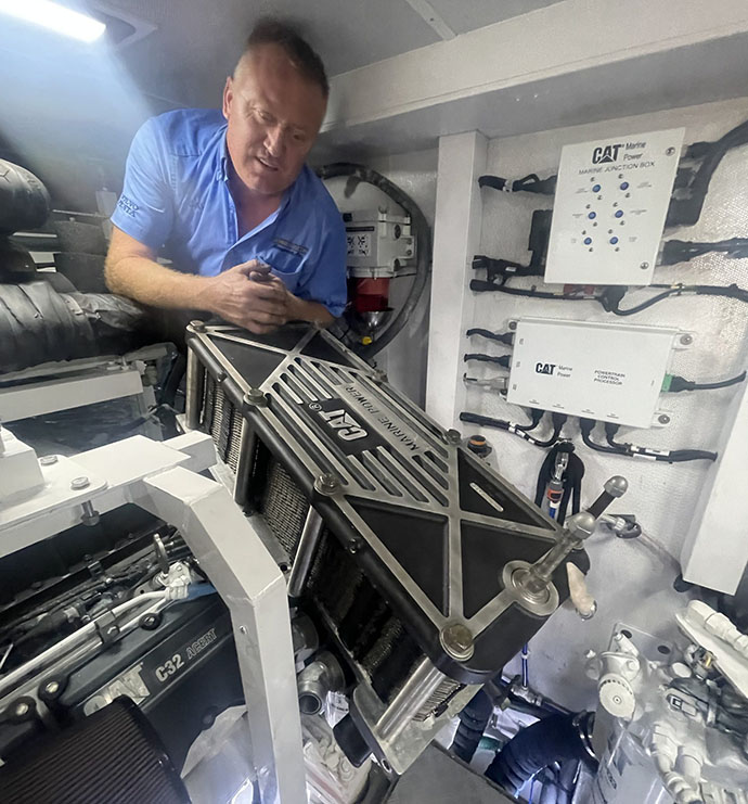 Technician handling a Cat marine heat exchanger beside a C32 diesel engine in a boat engine room.