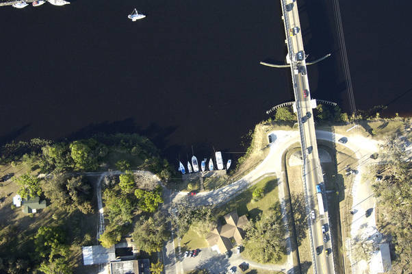 Aerial view of moored sailboats and a boat launch beside a bridge over a dark river.