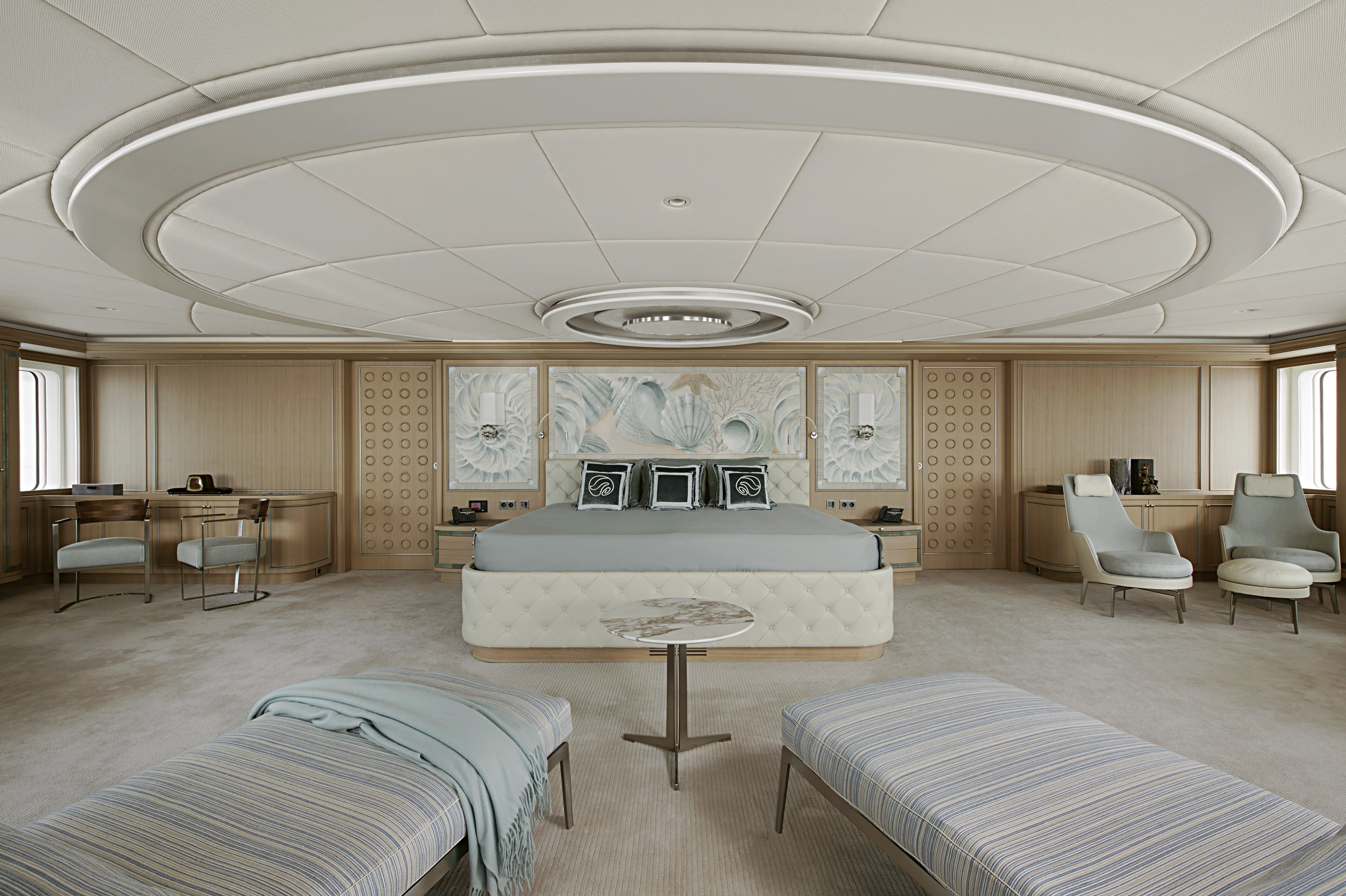 Master stateroom interior with a tufted white leather bed, striped upholstered lounges and shell-themed artwork aboard a luxury yacht.