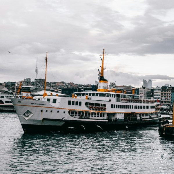 White double-deck passenger ferry with orange trim moored on the water in front of a hillside city waterfront.