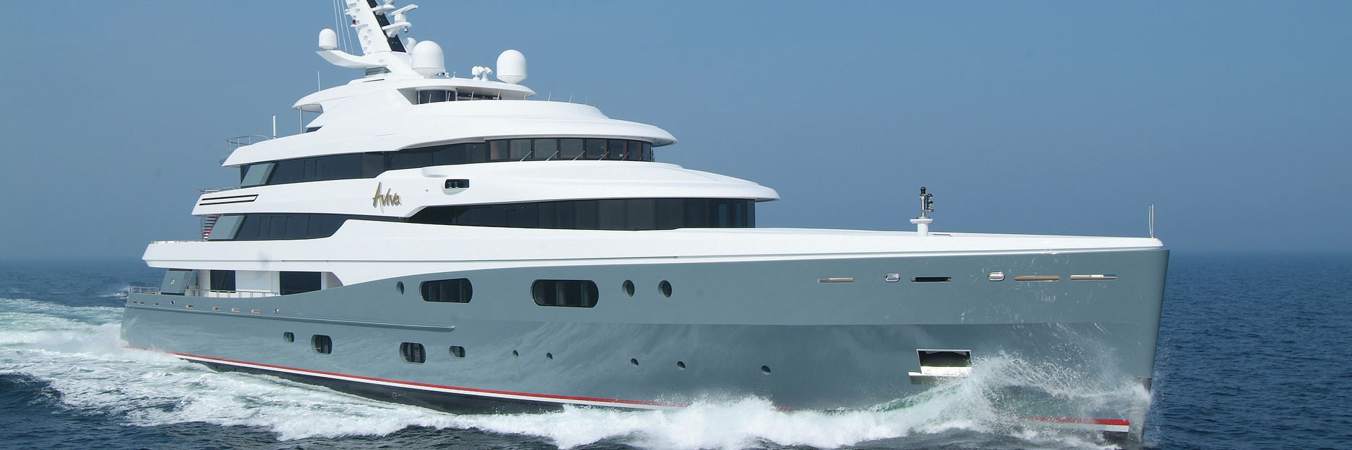 Motor yacht Aviva cruising at speed across open blue water, with a white superstructure, radar domes and a grey hull with red boot stripe.