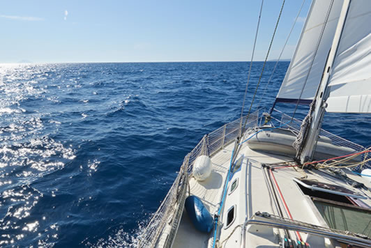 Sailboat deck with white sails and rigging cruising across open blue water on a sunny day.