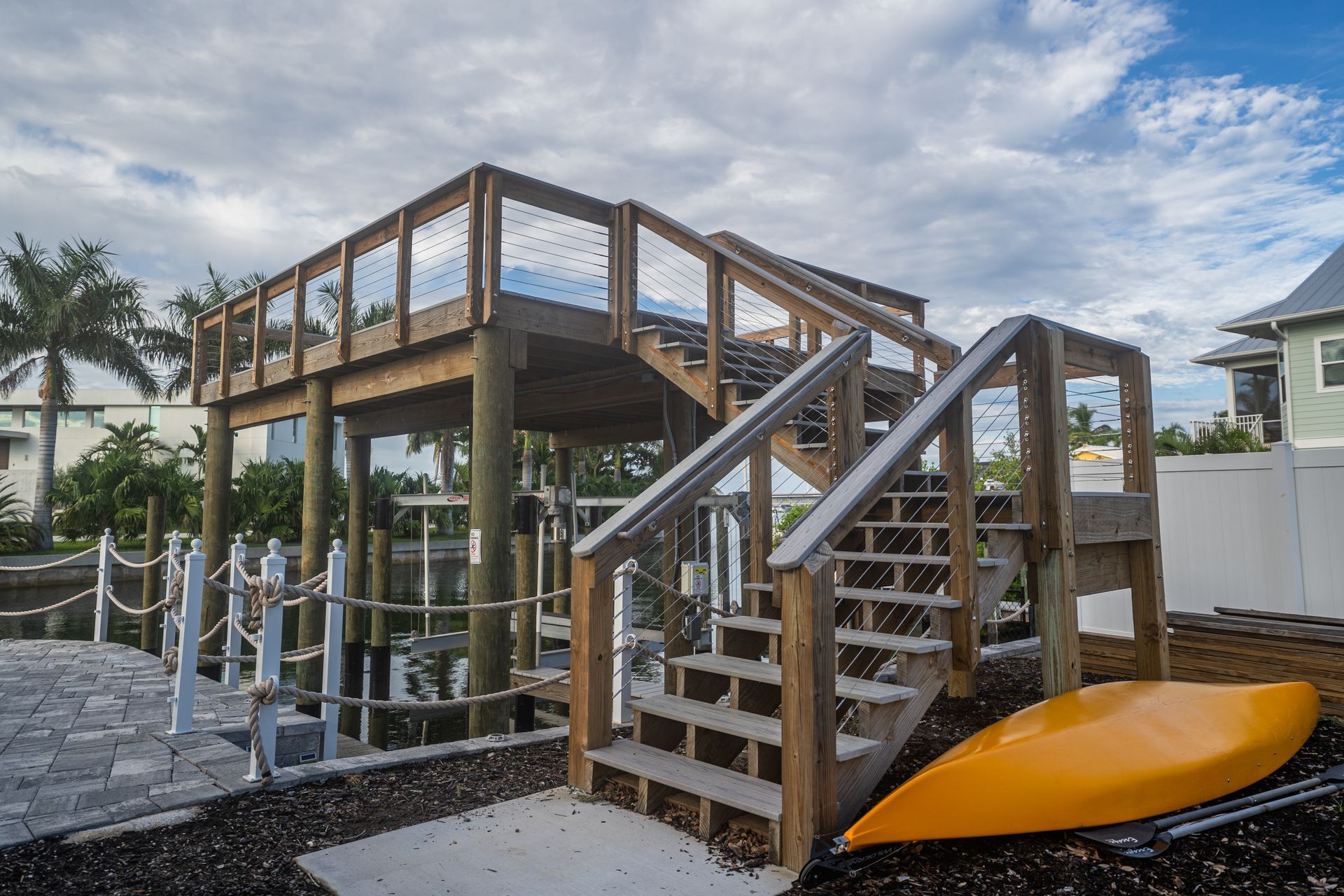Wooden elevated dock with cable railings, stairs and a boat lift over a canal, with an orange kayak resting on the shore.