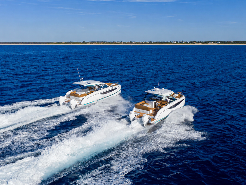 Two white motor yachts underway at speed on open blue water with a distant sandy shoreline, shown in an aerial photograph.