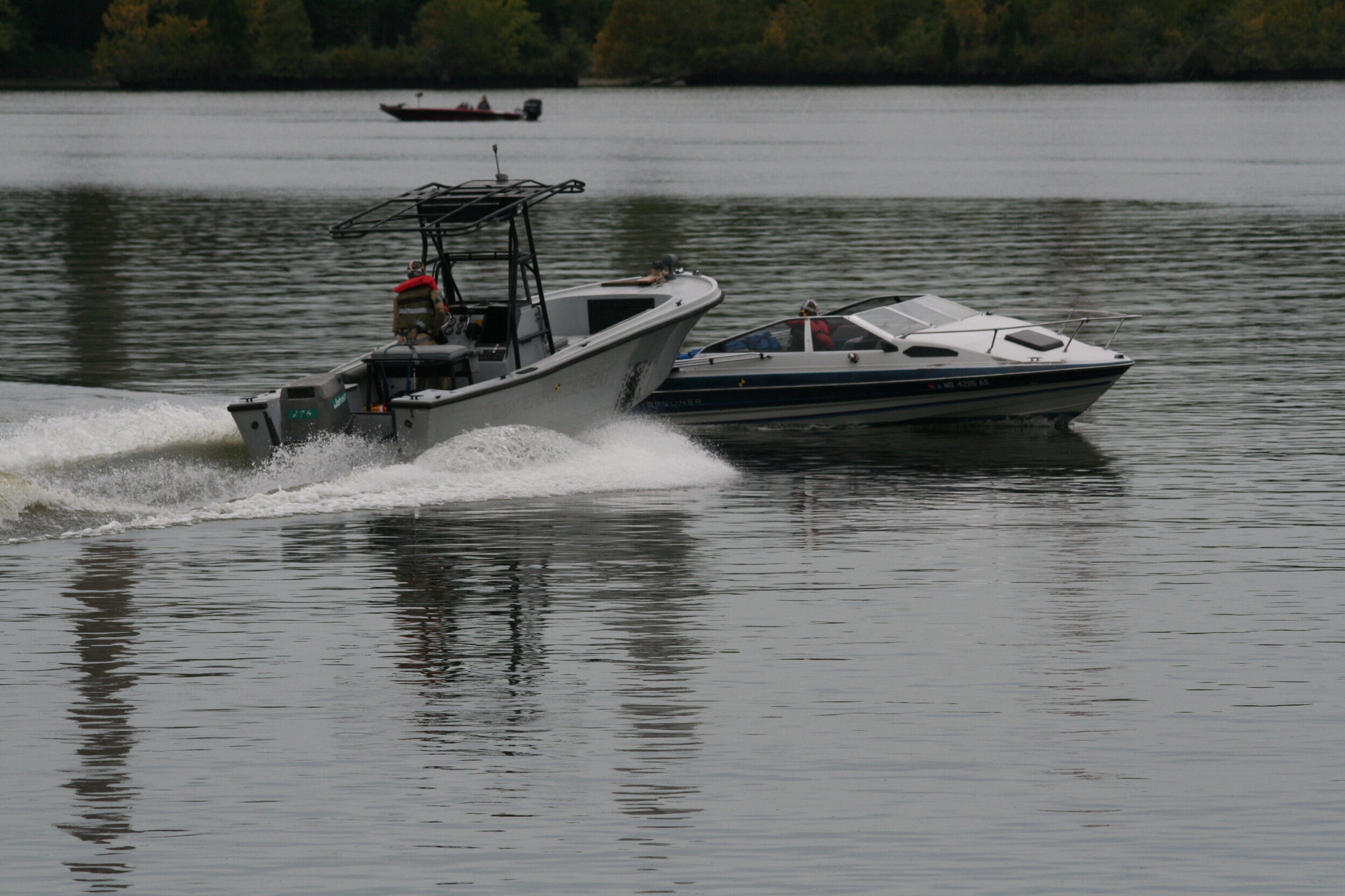 Gray center-console patrol boat with a white Bayliner cabin cruiser in tow on a river, with a red fishing boat in the background.