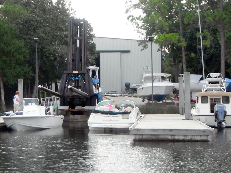 Travel lift hoisting a boat at a boatyard on the water, with docked boats and a storage building in the background.