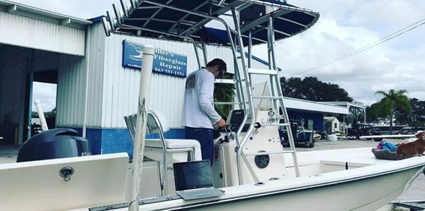 Man at the helm of a center-console boat with a T-top at Ike's Fiberglass Repair, a boatyard shop with a sign reading 863-551-1551.