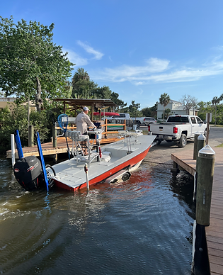 Flats boat with an outboard motor being backed down a launch ramp by a white pickup truck along a residential canal with a wooden dock.