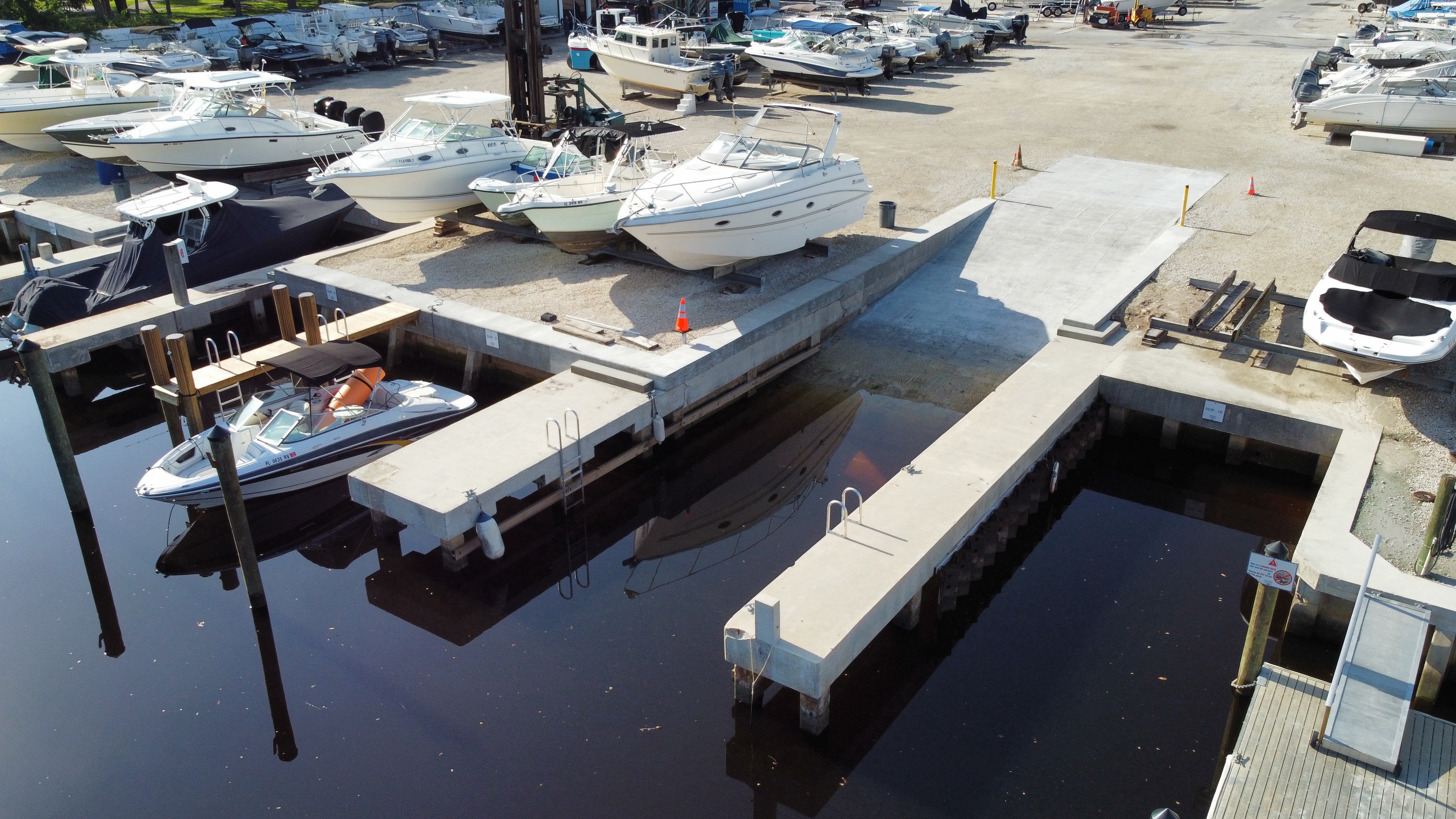 Aerial view of a concrete boat ramp, floating docks, and boats stored on the gravel yard at a boatyard, location unknown.
