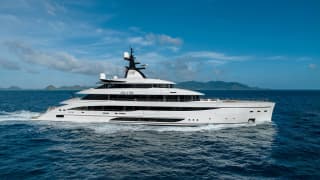 Large white motor yacht underway on open blue water with a hilly tropical island in the background.