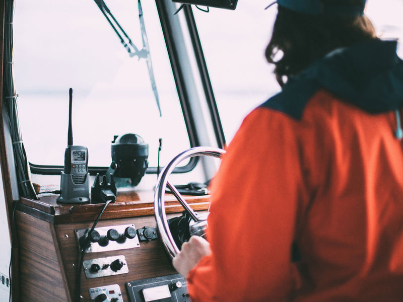 Person in an orange jacket steering a boat from the wheelhouse helm, with a VHF radio and control panel in view.