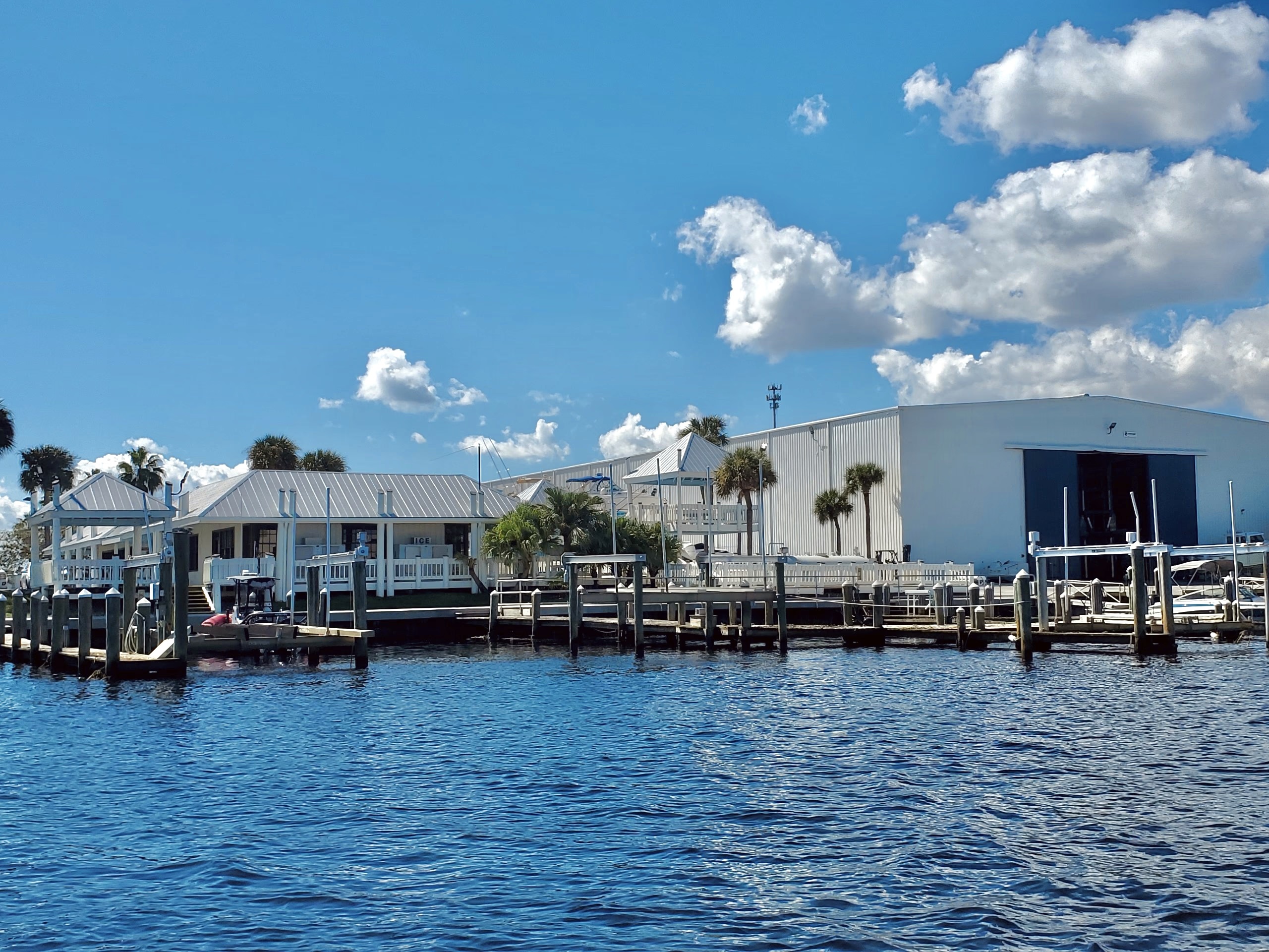 Waterfront marina with boat lifts, docks, and a large white boat storage building along the water.