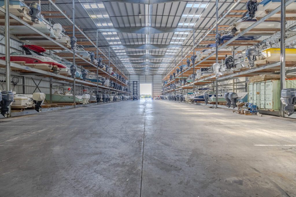 Interior of an indoor boat storage facility with powerboats stacked on multi-level racks and outboard motors lining the aisles.