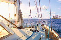 Sailboat deck with rigging and lines overlooking calm water and a small wooded island at sunset.