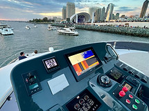 Boat helm with a chartplotter screen and view of boats in a harbor with a city skyline at dusk.