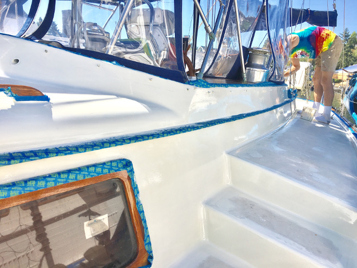 Freshly coated white sailboat deck and hull trim under blue masking tape, with a worker in a tie-dye shirt at the cockpit.