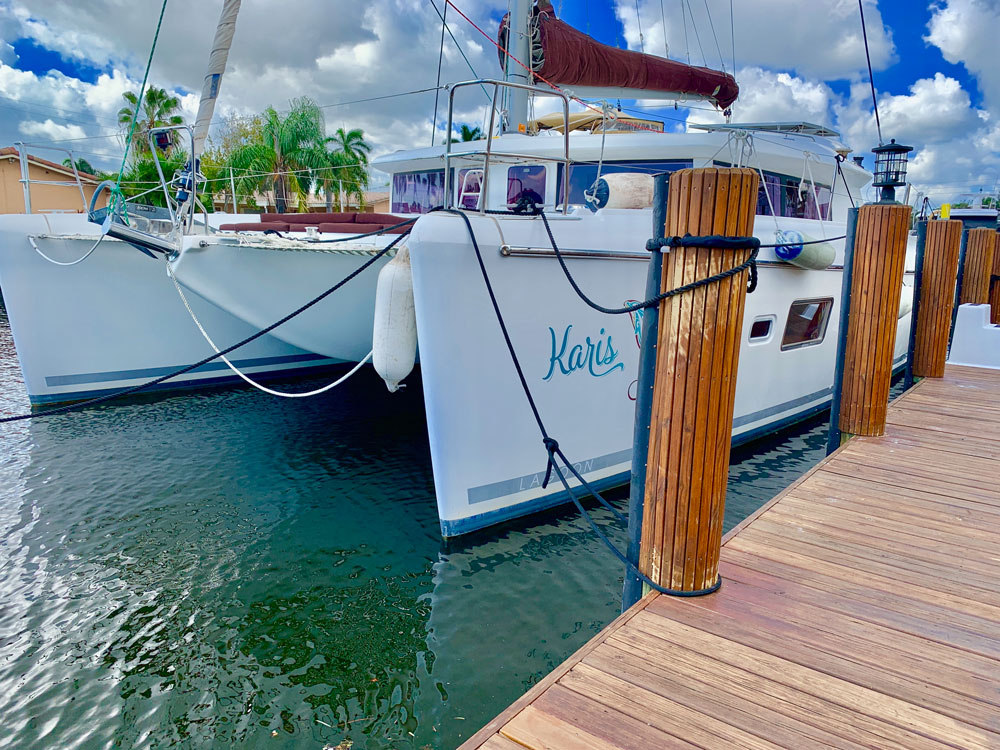 Catamaran named Karis tied up at a wooden dock with fenders and dock lines, with palm trees along the canal.