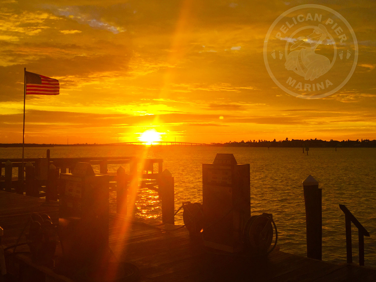 Sunset view from the dock at Pelican Pier Marina, with dock pedestals, hose reels, and a bridge across the water.
