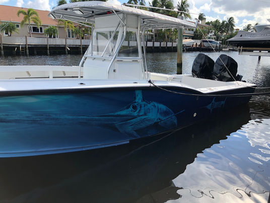 Center console boat with a blue marlin hull wrap docked in a canal, with palm trees and waterfront homes behind it.