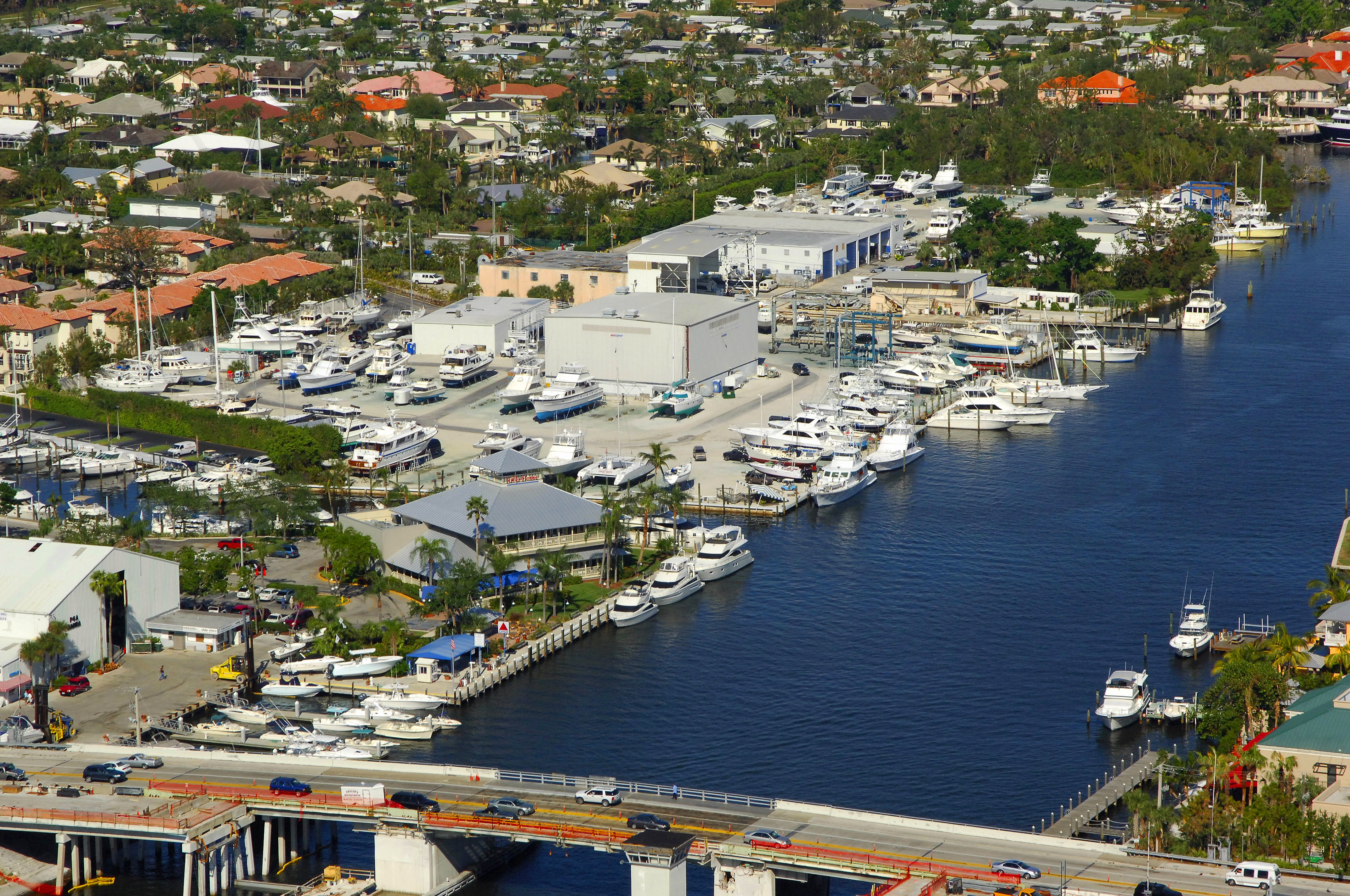 Aerial view of a full-service boatyard with boats on hard stands, dry-storage buildings, and yachts docked along a waterway beside a bridge.