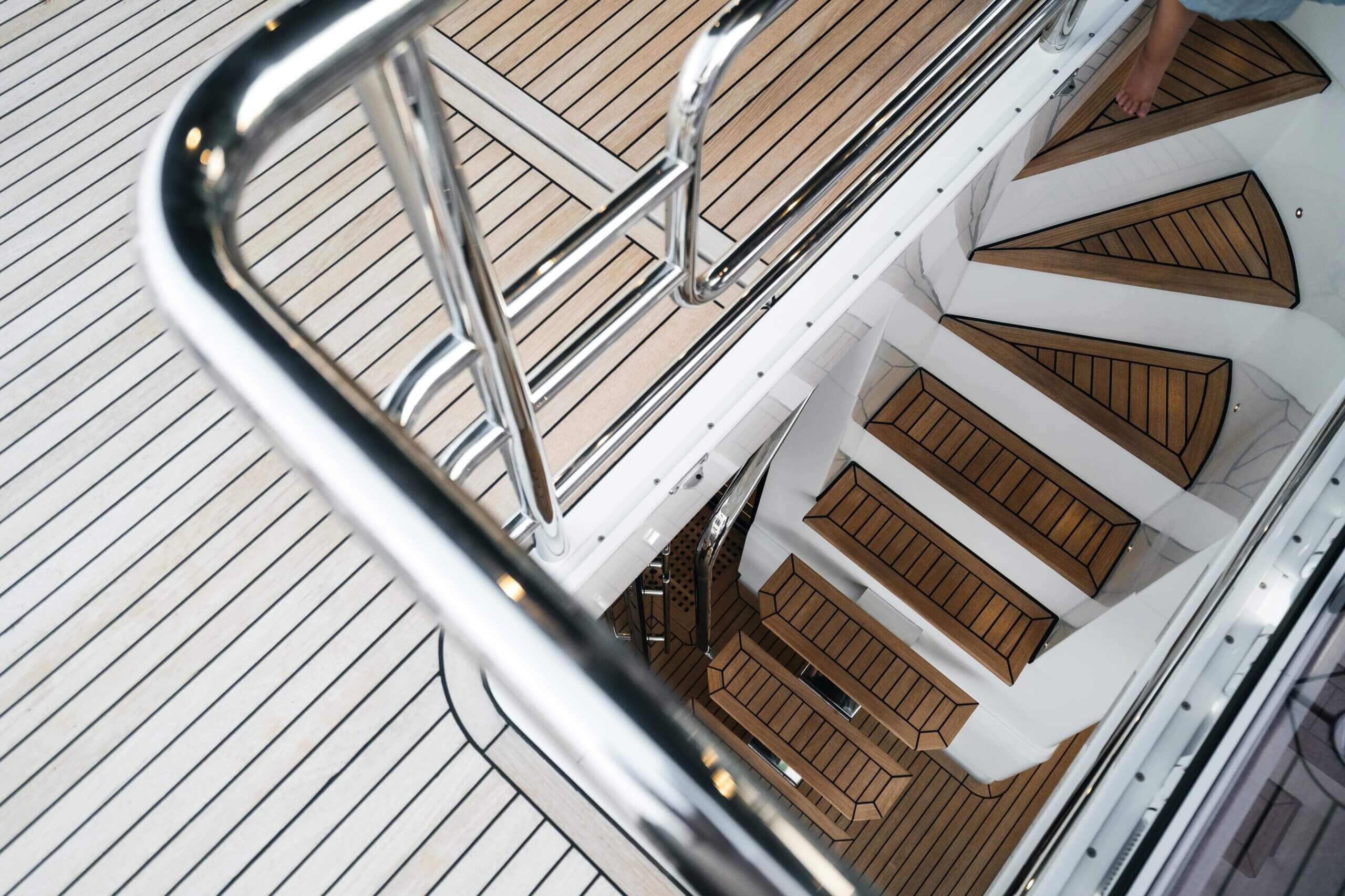 Teak and stainless steel companionway stairs on a yacht deck, shown from above with a person's bare feet on the top step.