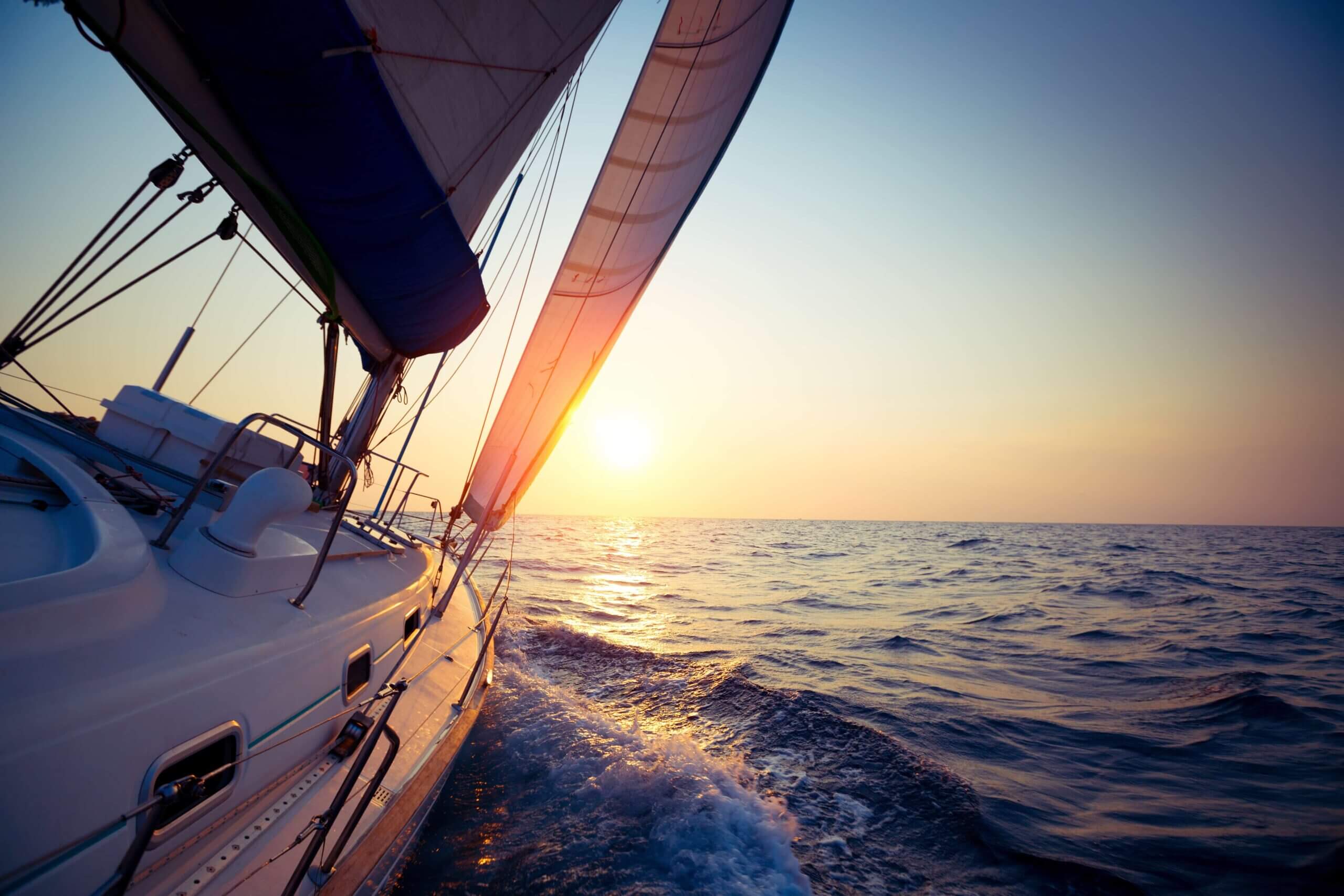 Sailboat under sail at sunset on open water, with its deck rigging and wake visible.
