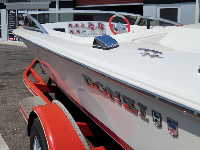 Vintage red-and-white Donzi speedboat with a red steering wheel and chrome fittings on a red trailer, parked in front of a full-service marine shop.