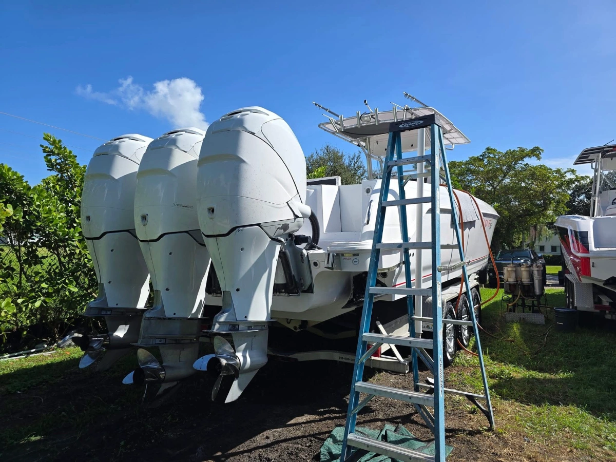 Center console boat with three white outboard engines on a trailer, with a ladder and service hoses set up on grass, likely a boat service yard in Florida.