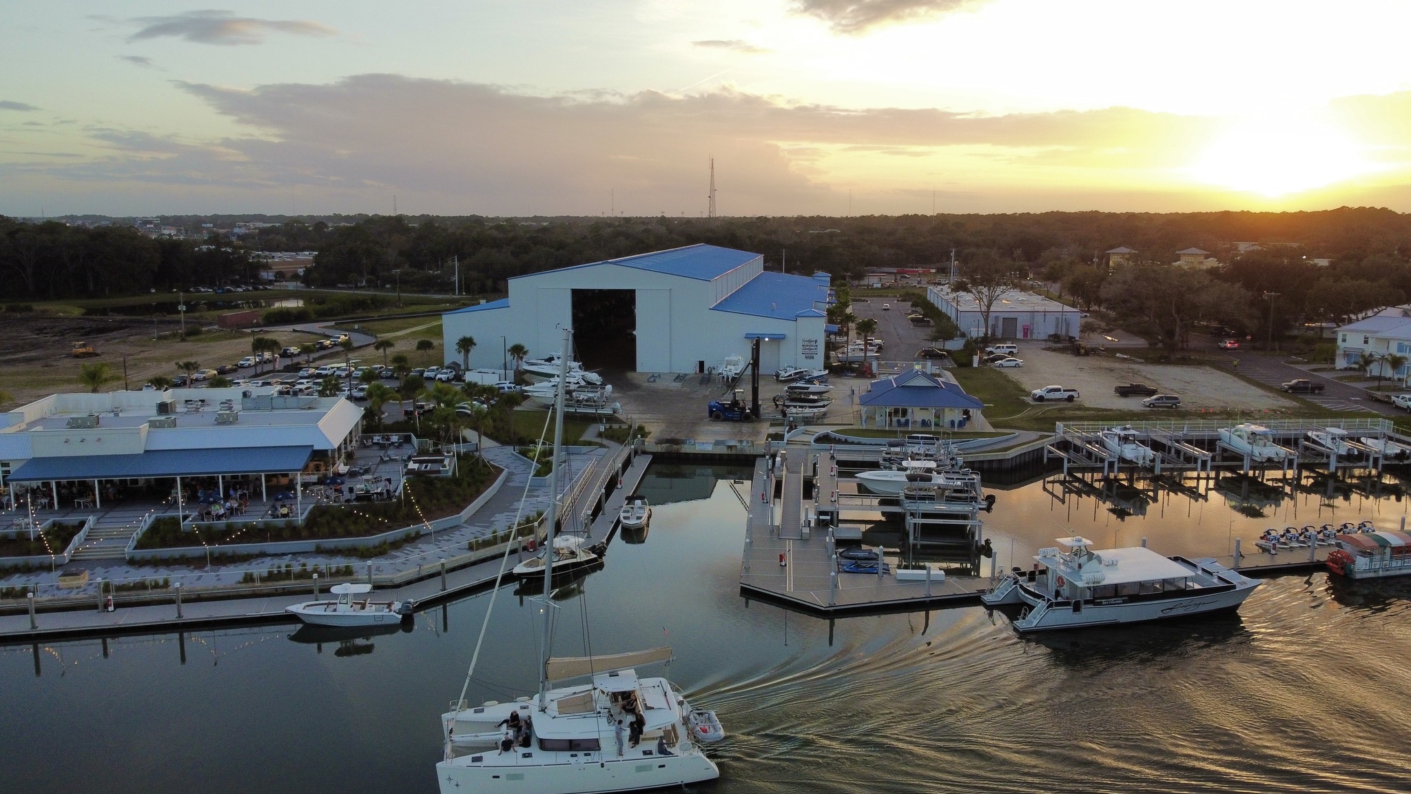 Aerial view of a marina with a large blue-roofed boat storage building, docked powerboats, and a catamaran leaving the basin at sunset.