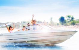 Friends waving from a speeding motorboat on open water near a sandy shoreline.