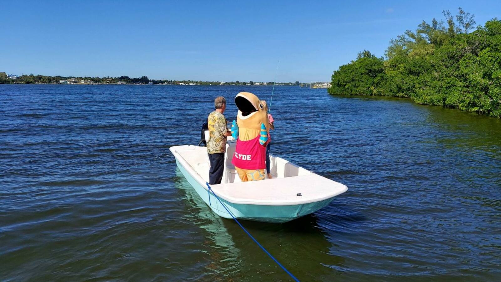 Small white and teal skiff carrying two people and a Clyde fish mascot on a calm waterway lined with mangroves.