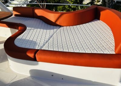 Orange and white quilted marine lounge seating on a boat deck, with a round table at the edge and palm foliage in the background.