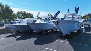 White center-console boats lined up in a parking lot at a boat dealership, with trees and blue sky behind them.