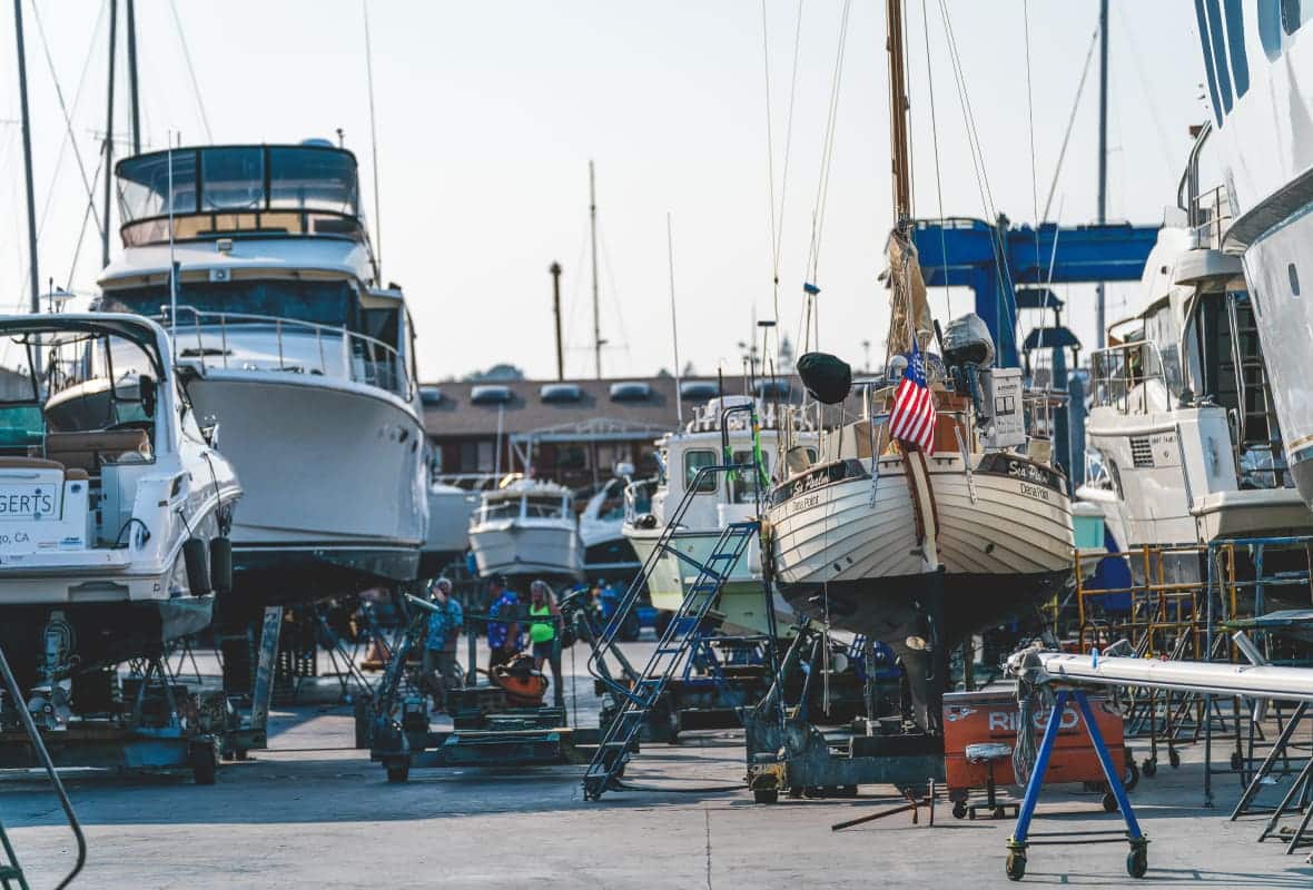 Yachts and a sailboat hauled out on stands for maintenance at a boatyard, likely San Diego, CA, business name not clearly visible.