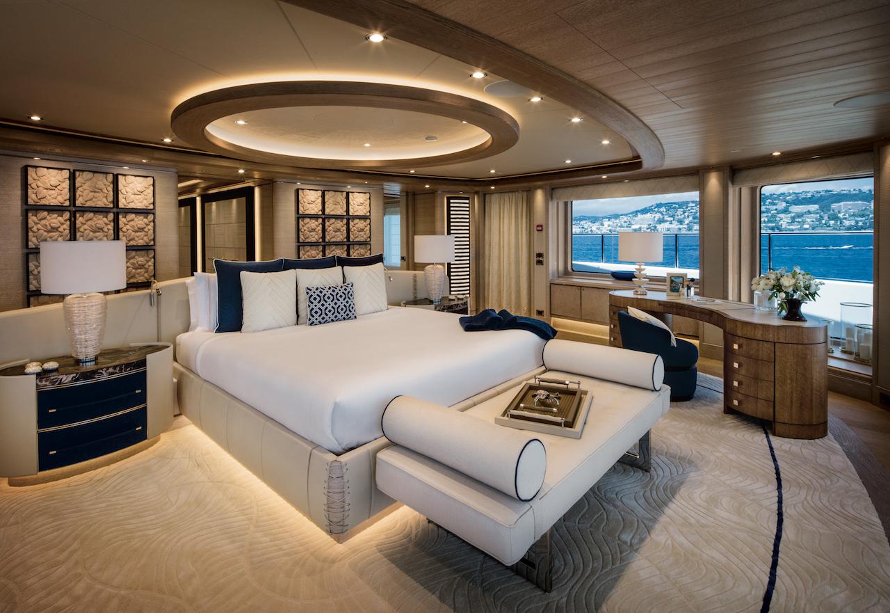 Master stateroom interior of a luxury motor yacht with a king bed, cream upholstery, and windows overlooking the water and a hillside town.