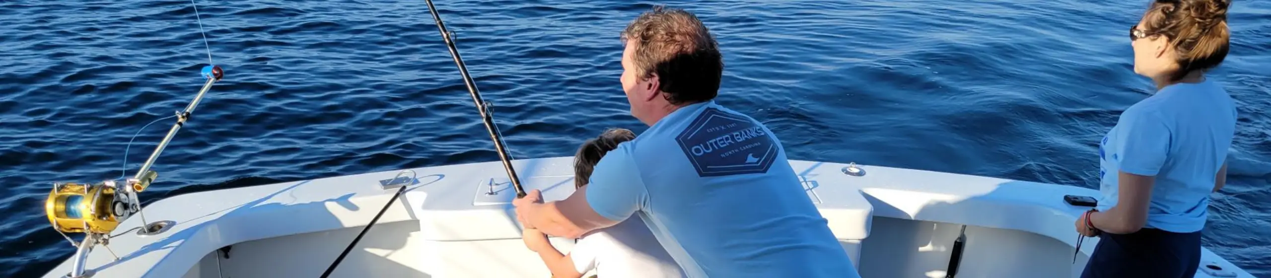 Two people fishing from the bow of a white sportfishing boat on open water, with a man in an Outer Banks North Carolina shirt holding a rod, likely offshore near the Outer Banks.