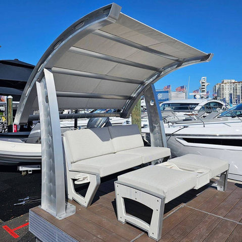Curved aluminum dockside canopy with cushioned bench seating and ottoman displayed among boats at a marina dealership.
