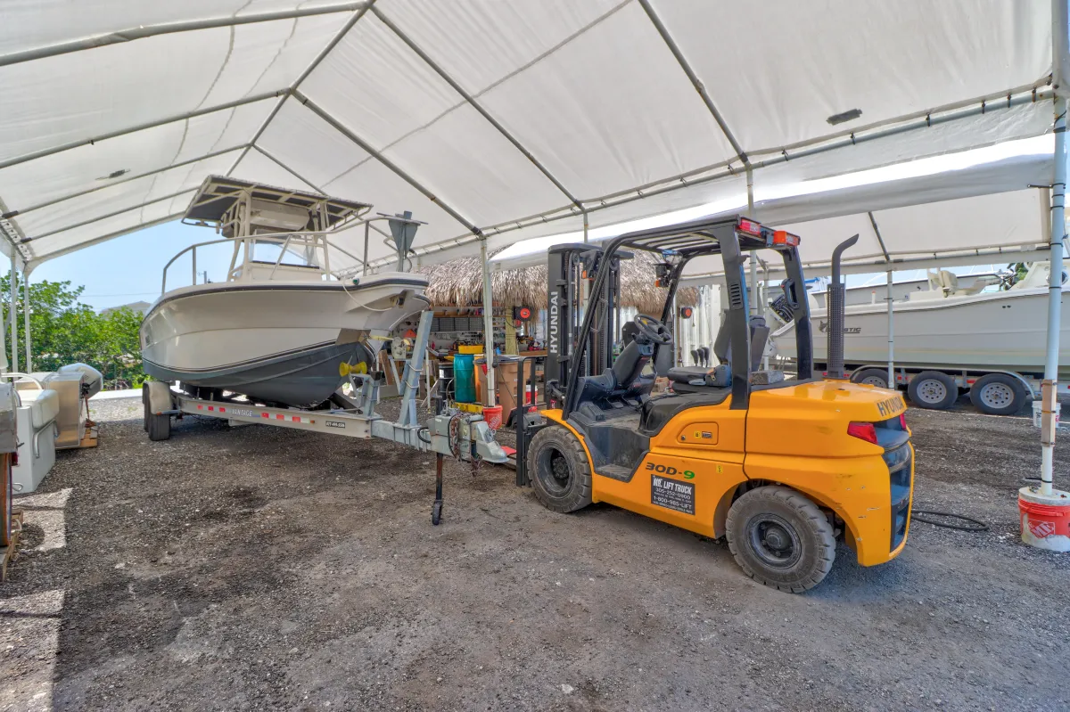 Center console boat on a trailer beside a Hyundai forklift under a storage canopy at a boat yard, with another boat stored nearby.
