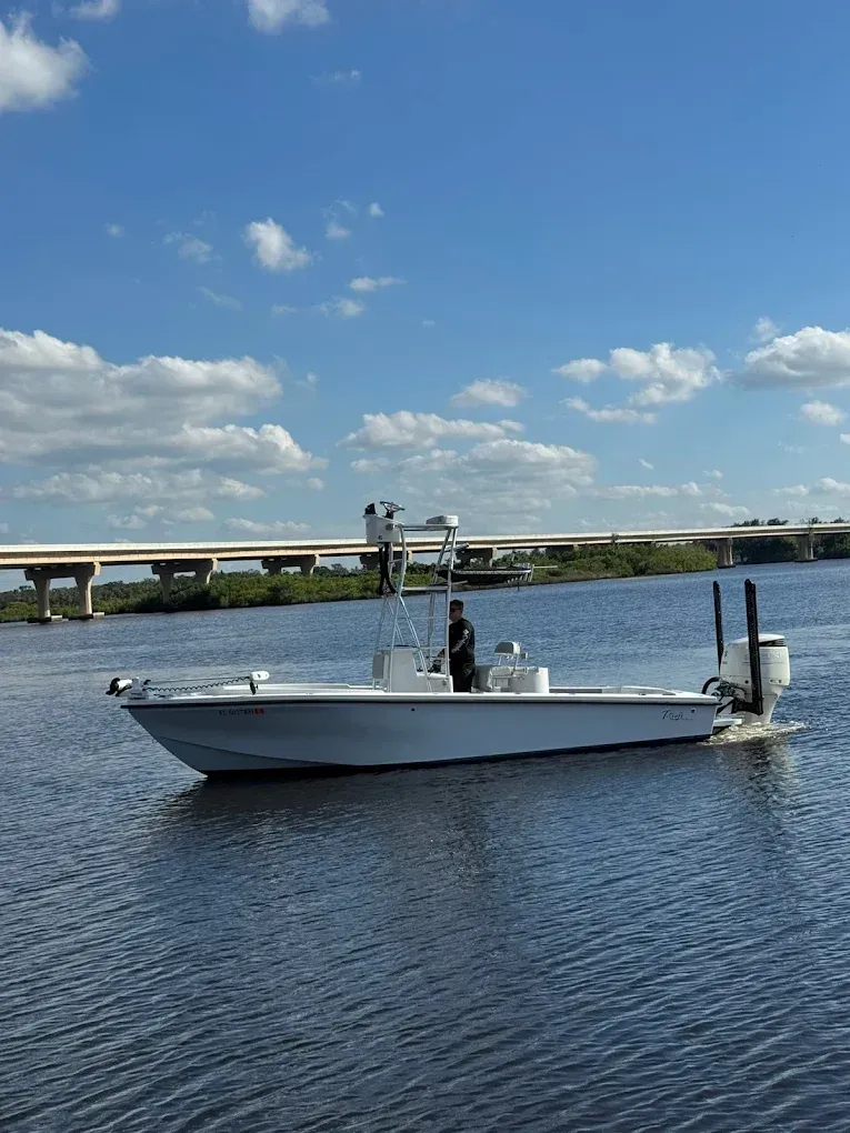 White flats fishing boat with a poling platform and twin outboards cruising a river under a highway bridge.