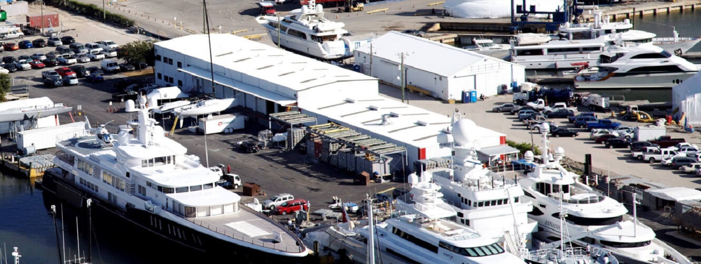 Superyachts docked beside large white repair sheds at a boatyard, likely a superyacht refit facility.