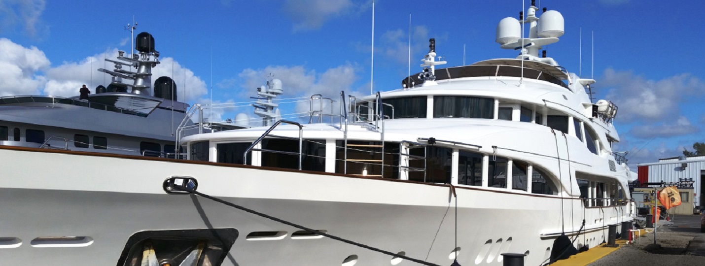 Two white superyachts tied up at a dock in a boatyard, with a yellow-edged pier and industrial buildings behind them.