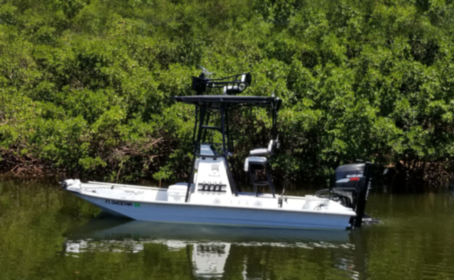 White center-console flats boat with a T-top and Suzuki outboard motor, anchored in calm water beside a mangrove shoreline.