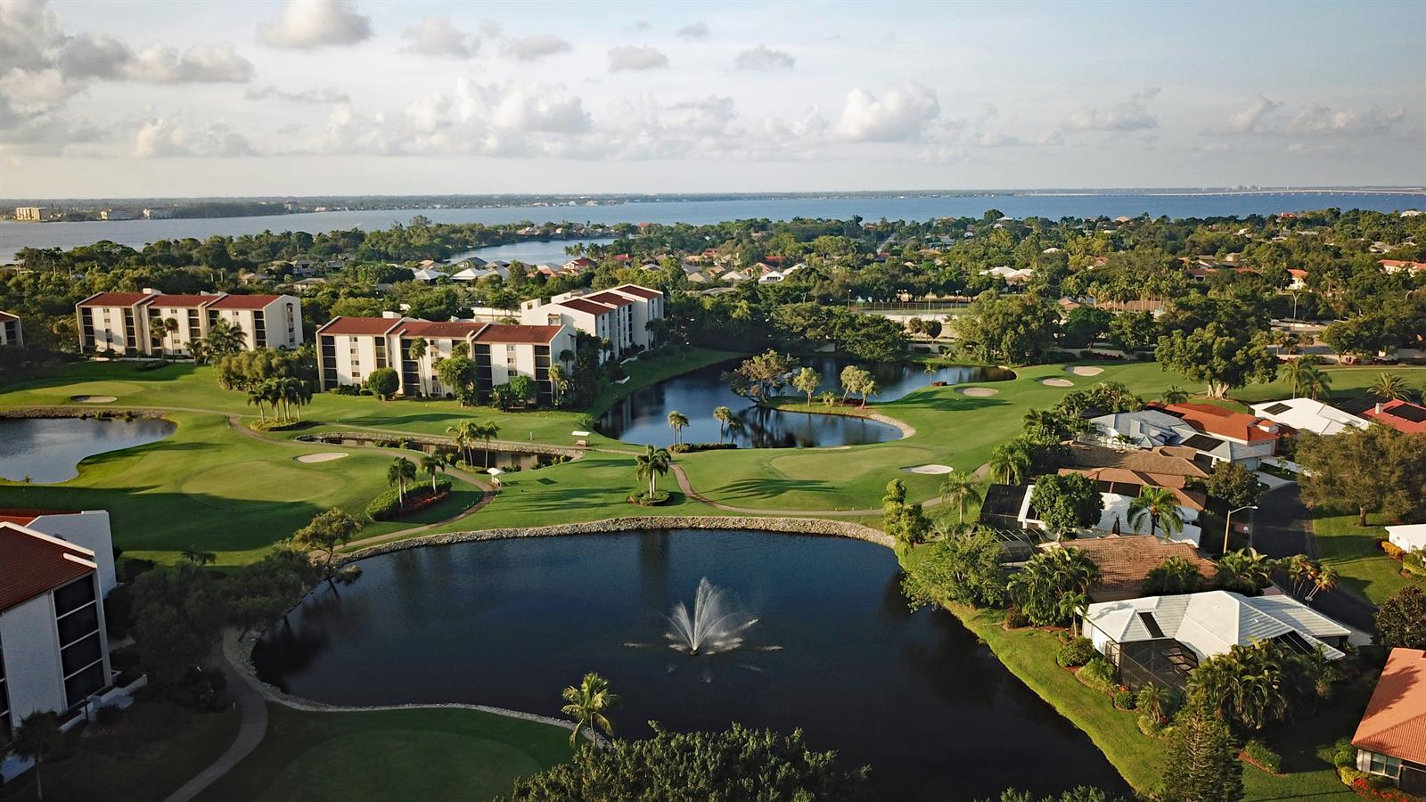 Aerial view of a golf course community with condominium buildings, a fountain-lit lake, and a bay in the distance, location unidentified.