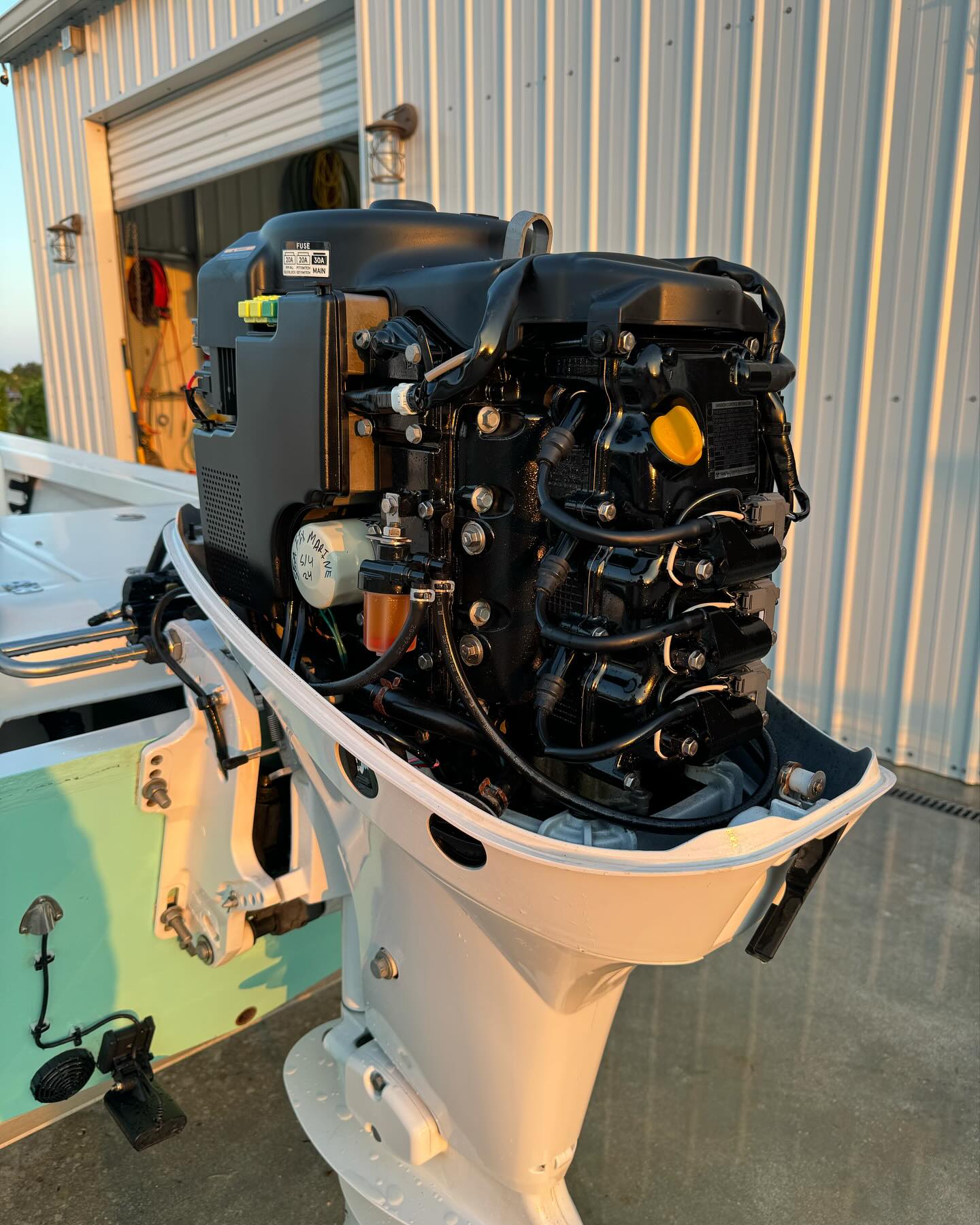 Outboard motor mounted on a white transom, with its cowling off and engine components exposed, set outside a metal-sided shop.