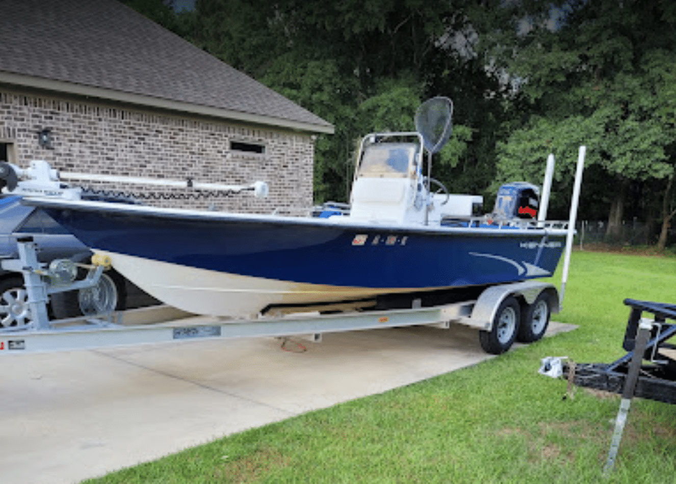 Blue center-console fishing boat with a white console and outboard motor, parked on a double-axle aluminum boat trailer in a driveway beside a brick house.